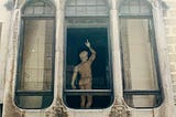 An outside window view of a statue of a naked man, from behind. He is looking up and pointing.