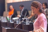 A woman stands behind a podium looking down at her notes while several people in the background sit at tables covered in water bottles.