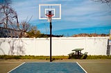 Image shows a lone basketball hoop in front of a white fence, with a blue sky behind it.