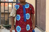 Young brown boy wearing African print and standing in front of a brown door.