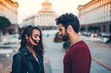 A woman and a man stand together in a downtown area, looking sad. The woman is dark-haired, in a black leather jacket, looking down in regret. The man is dark-haired as well with a short beard, and a long sleeved maroon shirt, looking at her in hurt.