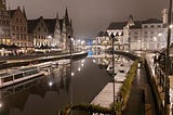 A view of the river Liei in Ghent taken from Grasbrug in the evening. A canal boat and several smaller boats sit on the water. People sit under umbrellas outside bars on the left of the river which is lit by lampposts on both sides. Sint-Michielsbrug is visible in the distance.