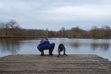 A woman and her dog, both crouched on a wooden dock over a lake, both leaning slightly to the left in sink.