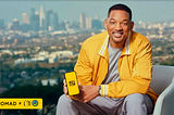 Will Smith sorrindo, sentado em uma poltrona clara, vestindo jaqueta amarela, segurando um smartphone com tela amarela exibindo um logotipo estilizado. Ao fundo, uma vista ampla de uma cidade com prédios ao longe. No canto inferior esquerdo, há uma faixa amarela com os logotipos “Nomad” e “L3TI”.