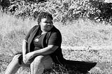 A photo of Krystal Toney: she is a Black woman sitting on grass with trees in the background and looking to her left while smiling