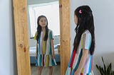 Little girl with long, black hair in striped dress, looking at herself in the mirror