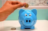 A hand dropping a coin into a piggybank; you can also save money as a writer by getting free writing lessons