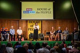 A stage with a woman speaking surrounded by people sat on chairs, on the stage and in the audience