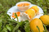 On a white-plate, a cylindrical rice cake with yellow color is served with visible mango cut in between them. Around it, a bowl of mango puree, cuts of mangoes, and two mangoes are placed.