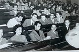 Black and white photo of dark haired students in an auditorium posing for a photo