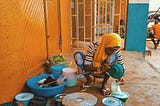 Woman in Bida (Niger State), Nigeria on a low seat serving food into a plastic bag. She is surrounded by pots and bowls, wearing an orange head covering in front of an orange-painted, closed mattress shop