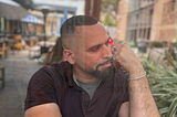 International best sellling author christopher knight lopez sitting in a park with a phone to his ear looking away from the camera.