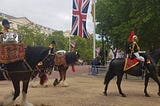 Three horses walking past, two of which are carrying silver kettle drums with their musician riders; one in front is carrying a soldier.