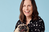 Image shows a lady with long dark hair, smiling and cuddling a pug dog