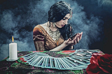 A beautiful tarot reader is surrounded by a candle, a red flower, and tarot cards fanned out on a table in front of her. She has one card in her palm, examining it for it’s meaning.