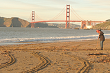 View of the Golden Gate Bridge from Baker Beach at sunset with woman raking sand in foreground