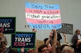 Posters are held above trans people and their allies reading “Being Trans is NOT a choice Protect gender affirming healthcare we will NEVER give up” and “Protect Trans Kids” and “Actual Floridian”