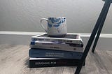 A blue and white coffee mug sits atop a stack of 5 books, on the floor, against a wall. Next to them are the legs of a lamp.