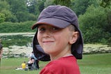 A young boy around 10 years old. He has a red t-shirt and a blue cap with a sun guard protecting his neck. He is looking at the camera and smiling. People are in the background admiring a lake, and the boy is surrounded by trees.