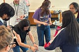 Eight people standing and looking down at a desk with a very small battery powered robot; smiles abound, and a snack cart in the background.