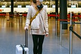 Woman wearing a mask looking up in an airport terminal with her rolling carry-on bag.