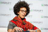 Timnit wearing a red patterned scarf and a grey sweater, speaking on stage.