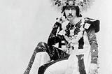 Paget c. 1900, photographed by John Wickens. He’s wearing a winged headdress, and an extraordinary, elaborate skirted costume and tights