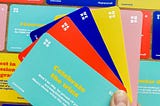 A thumb holds a selection of brightly coloured PR prompt cards in green, red, blue, yellow and pink on top of a backdrop of cards.