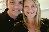 Close-up photo of the Author with her husband posing for a selfie in front of a white wall. The author has shoulder-length blonde hair and is wearing a black dress. The author’s husband has short brown hair and is wearing a black shirt.