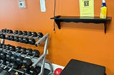 A gym area with a bright orange wall featuring a set of black dumbbells on a silver rack, an ab roller, a red medicine ball, and a stack of black exercise mats. Note that the wall includes a small black shelf holding cleaning supplies, alongside signs reminding users to wipe down machines and indicating first aid locations.