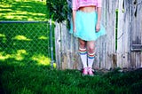 On an outside lawn, leaning against a fence which is 1/2 chain link (left side) and 1/2 wooden boards (right side) leans a caucasian person. Torso to feet are visible, head is not. Pink half shirt, bare midriff, turquoise poofy skirt, and rainbow kneesocks leading to pink converse sneakers are worn.