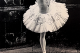 A black and white photo of a ballerina standing a top a metal bucket stand, in a lace tutu. The black tiles in the background reflect the white of the ballerinas tutu