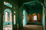 A view into several rooms of a beautiful Moroccan space, with sea-foam/teal walls, high contrast tile flooring, ornately arched doorways, and sunlight pouring in through windows.