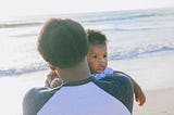 Photo of African American father holding infant son on his shoulder, facing the ocean with the father's back to the camera.