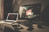 Laptop set on a desk next to a flat screen monitor, with keyboard, mouse, phone, and tablet, all scattered above the desk