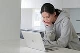 Young, Asian woman staring at a laptop in a minimalist kitchen.