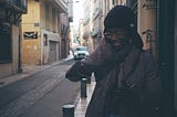 Toyin during their trip. They are wearing a coat and hat, holding a cig and grinning on a tight street in Perpignan, France