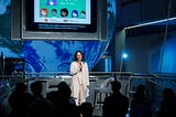 A woman in business casual clothes stands on a stage speaking to an audience at the “Day of AI” event, held on May 13, 2024, in a museum. Behind her is a large screen displaying graphics related to artificial intelligence and a giant globe.