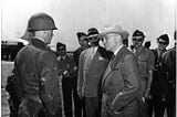 President Harry Truman stands in front of a group of soldiers on an airplane tarmac. He wears a brown suit, a white hat, and has his hands in his suit pockets. His head is turned to look at a soldier in a uniform and helmet, whose face we see in profile.