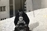 Black-and-white photo of young child with older man on sled, apartment buildings behind them
