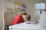 A woman with her forehead on her desk in front of her.