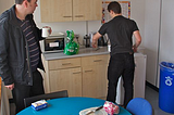 Two men in an office kitchen making coffee and tea