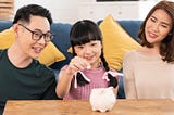 Two parents smiling with a child who is placing a coin in a piggybank.