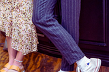 Photo of a teenage couple, with just their legs pictured. He’s wearing pinstripe dress pants and classic white high-top Converse. She’s wearing a summery dress and platform sandals, her toenails a light, turquoise blue. You can see that they’re holding hands.