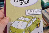 Photo of a hand Holding the book titled “Cyber Car 2010”. The cover has an illustration of a green sedan with the sign “cyber car has arrived” in the background. The book is by Daniel Pluck. Ilustrations by Duncan Scott
