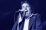 black and white photograph of a man in a suit jacket and collared shirt singing into a microphone with his eyes closed