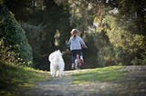 A girl rides a bicycle in a natural road followed by her little white dog