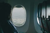 A woman, nearly silhouetted, young-ish, stares out the window of an airplane. Tones in photo are mostly greys and blues.