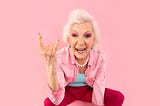 badass lady with grey hair, rockstar makeup, giving the devil’s horns with the fingers on her hand.
