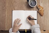 A woman writing in a notebook wearing silver rings. There is a croissant and a cup of coffee next to her.